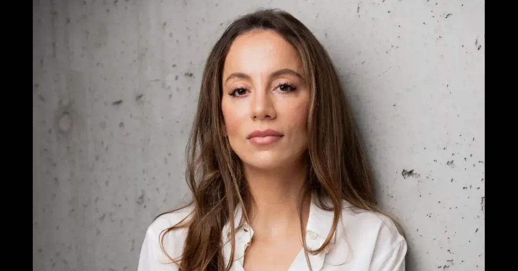 Mira Murati, with long brown hair and wearing a white blouse, stands confidently against a light gray concrete wall, looking directly at the camera.
