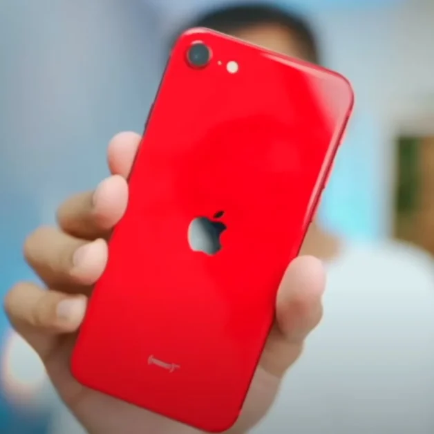 Person holding a red iPhone SE 4 (2025) showing rear camera and Apple logo.